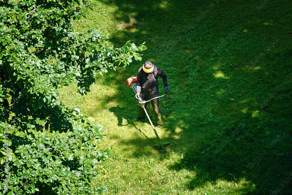 Worker trimming grass, aerial view. Worker mowing lawn in residential area, cutting and landscaping. Man mowing grass, municipal worker cutting grass and maintaining public lawns for neat appearance