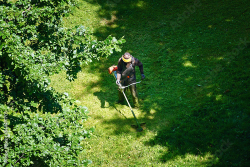 Obraz na płótnie Worker trimming grass, aerial view. Worker mowing lawn in residential area, cutting and landscaping. Man mowing grass, municipal worker cutting grass and maintaining public lawns for neat appearance w salonie