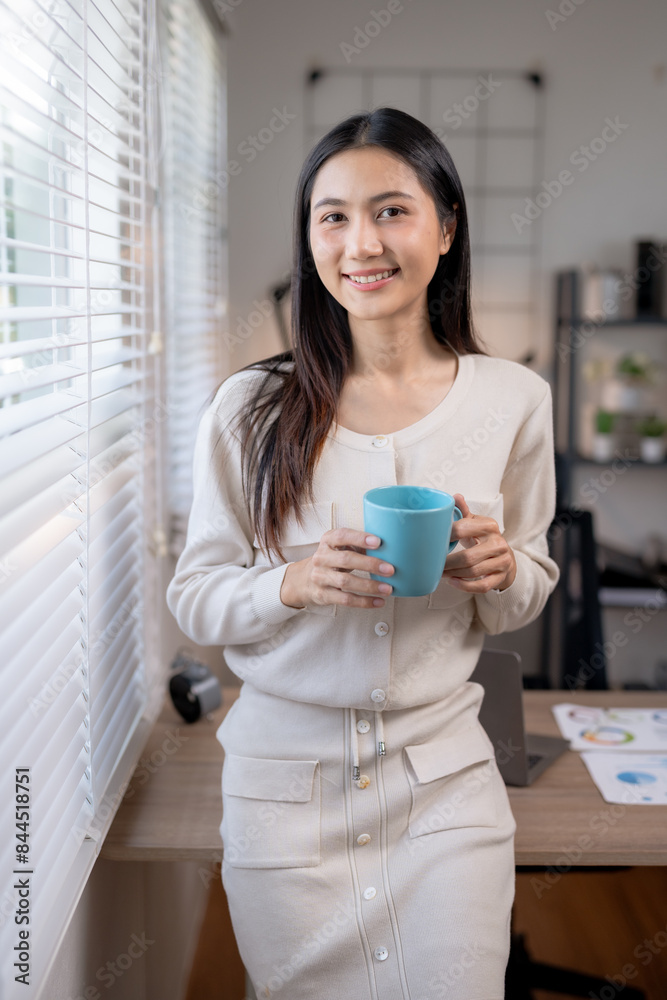 Businesswoman in white attire, holding a blue mug and smiling, standing by a window in a modern office setting. Businesswoman in white attire, holding a blue mug and smiling, standing by a window in a modern office setting.