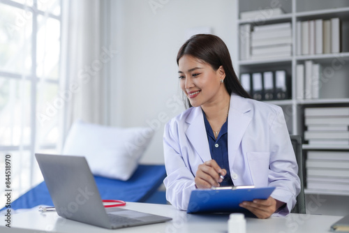 Female doctor working on a laptop while taking notes on a clipboard in a modern medical office. Healthcare professional in a clinical setting.