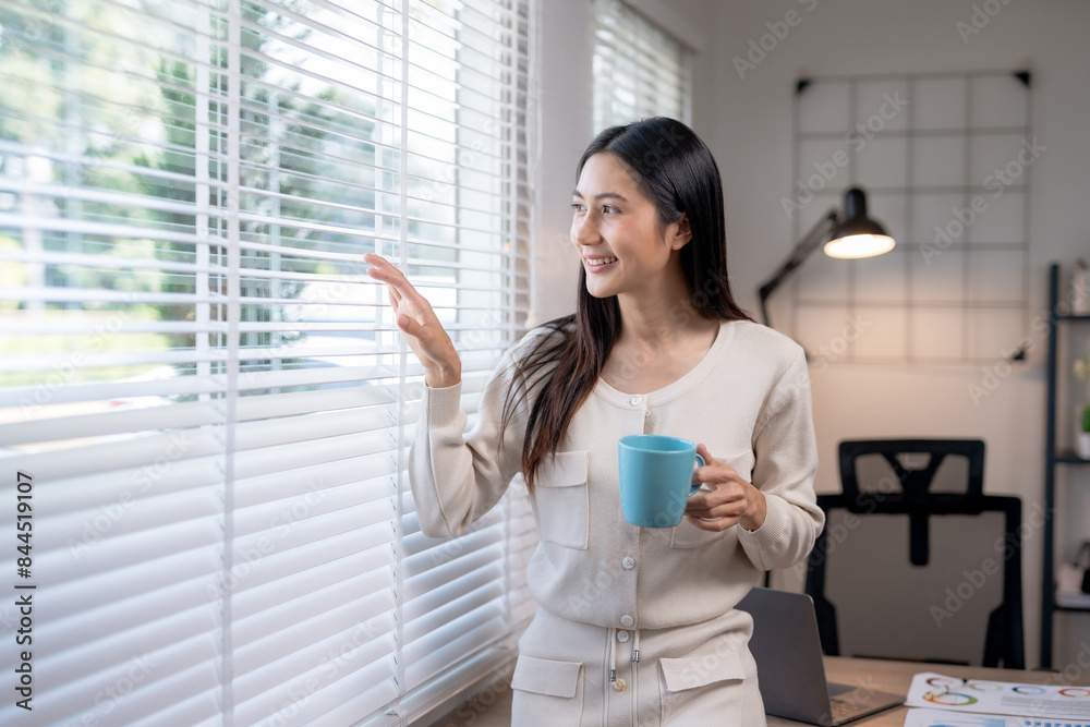 Smiling woman holding a blue cup while looking through window blinds in a modern home office with natural light and comfort. Smiling woman holding a blue cup while looking through window blinds in a modern home office with natural light and comfort.