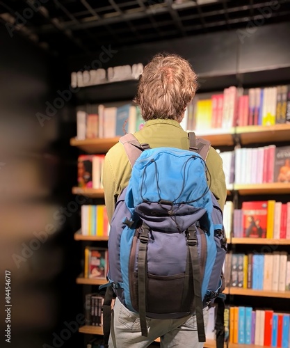 Young adult man,standing,seen from behind,with backpack on his shoulders,looking at a book to buy for the plane trip.Man from behind, standing,looking in the airport bookstore.Attending a bookshop. photo