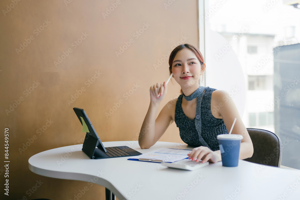 Obraz premium Young Professional Woman Working at Modern Office Desk with Tablet and Coffee, Smiling and Thinking, Bright Natural Light from Window, Business and Productivity Concept