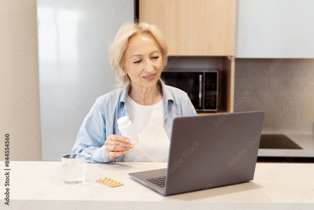 Senior Woman Holding Medication While Using Laptop In Kitchen, Elderly Care And Online Health Consultation Concept Senior Woman Holding Medication While Using Laptop In Kitchen, Elderly Care And Online Health Consultation Concept