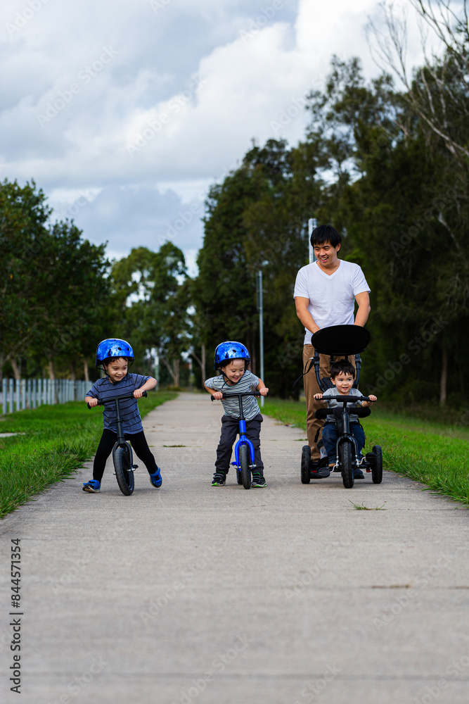 Chinese Australian dad on walk with his sons riding balance bikes on footpath Chinese Australian dad on walk with his sons riding balance bikes on footpath