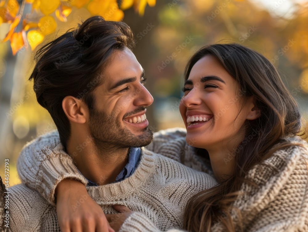 Obraz premium A happy couple in cozy sweaters embraces outdoors, both smiling brightly, with autumn leaves in the background.