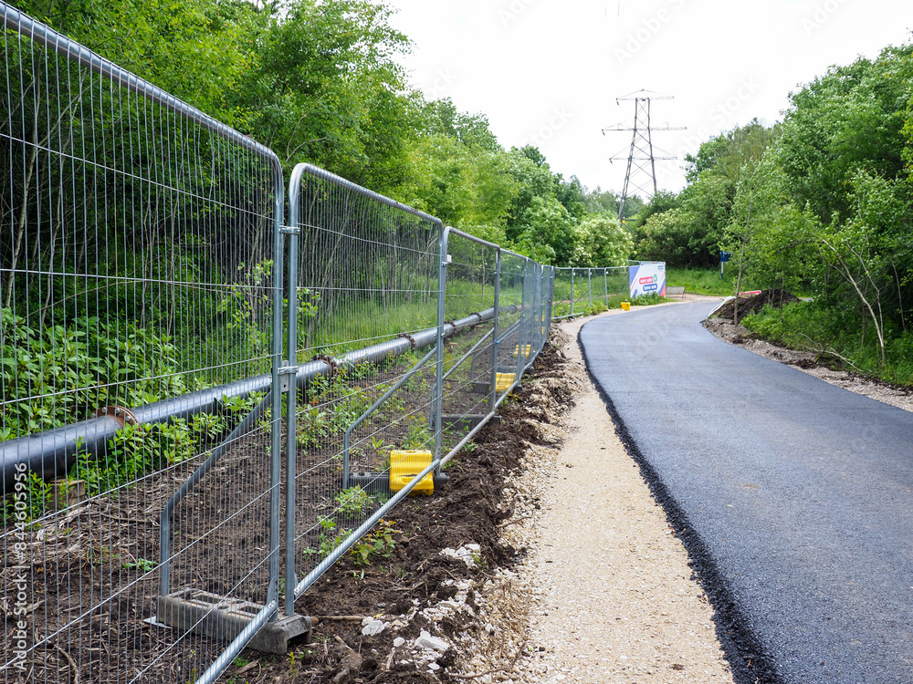 Freshly paved road alongside a construction site with safety barriers and lush green trees. Freshly paved road alongside a construction site with safety barriers and lush green trees.