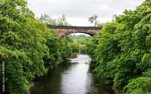 A stone bridge arches over a serene river Irwell Salford surrounded by lush greenery on a calm, cloudy day. photo