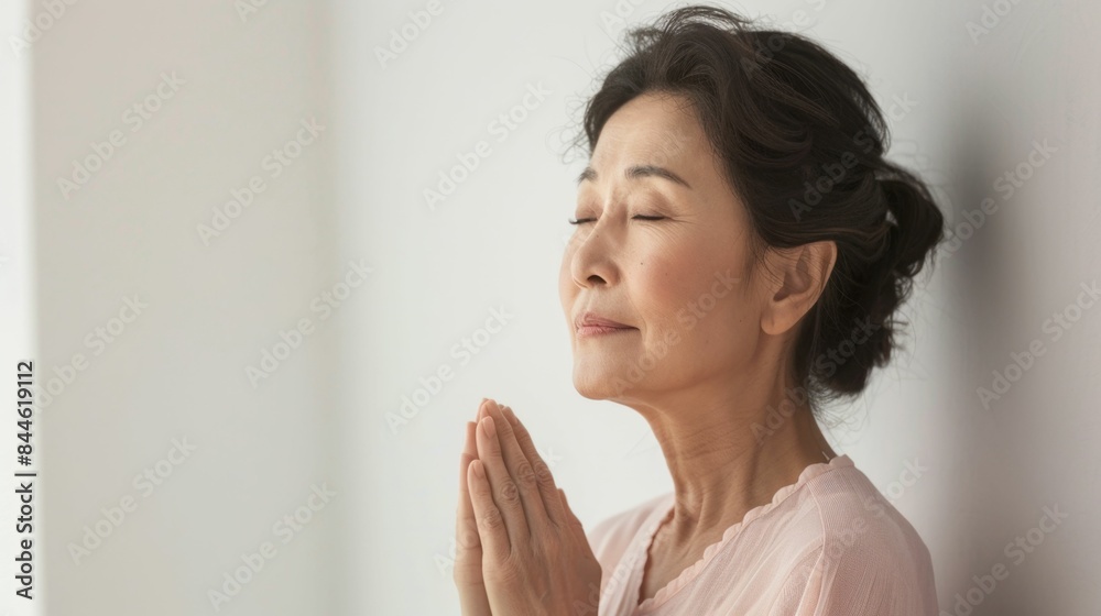 A serene woman with closed eyes hands clasped in prayer standing against a white wall. A serene woman with closed eyes hands clasped in prayer standing against a white wall.
