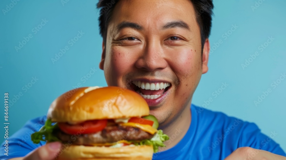 A man with a broad smile holding a large appetizing burger with a variety of toppings against a vibrant blue background. A man with a broad smile holding a large appetizing burger with a variety of toppings against a vibrant blue background.