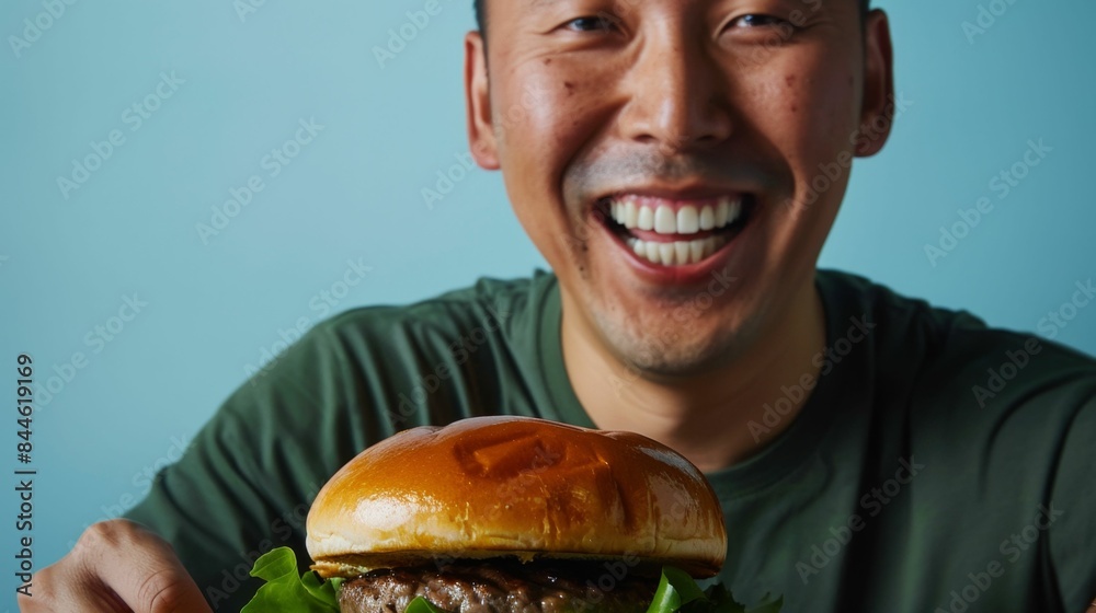A cheerful man in a green shirt holding a large appetizing burger with a bun that has a distinct grill mark. A cheerful man in a green shirt holding a large appetizing burger with a bun that has a distinct grill mark.