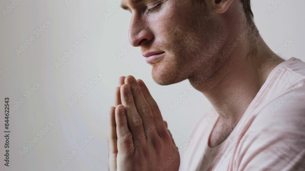 Fototapeta premium A man with a beard and closed eyes clasping his hands together in a gesture of prayer or meditation wearing a light-colored shirt against a neutral background.