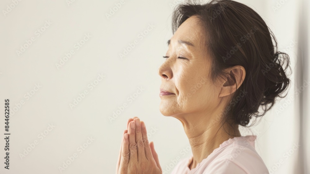 A serene elderly woman with closed eyes hands clasped in prayer standing against a plain white background. A serene elderly woman with closed eyes hands clasped in prayer standing against a plain white background.