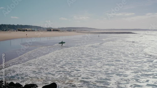 A lone surfer walks towards the ocean on a vast, empty beach in Portugal. Waves crash gently against the shore under a clear sky. Aerial view of man carrying surfing board and go into ocean 