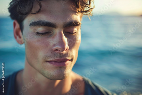 Close up of a handsome young man meditating, eyes closed, on the shore of a peaceful ocean.