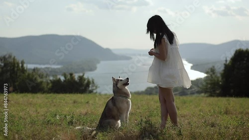 young brunette woman standing together with a siberian husky dog on a field with mountains and a river in the background in slow motion photo
