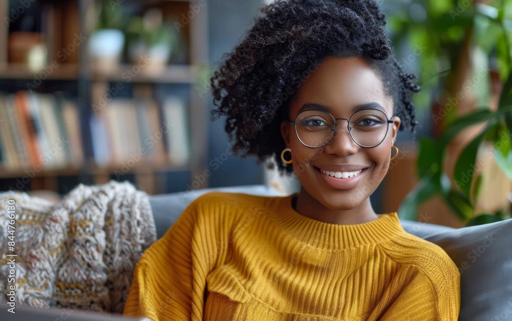 A woman with curly hair and glasses is sitting on a couch and smiling. She is wearing a yellow sweater and is surrounded by books A woman with curly hair and glasses is sitting on a couch and smiling. She is wearing a yellow sweater and is surrounded by books