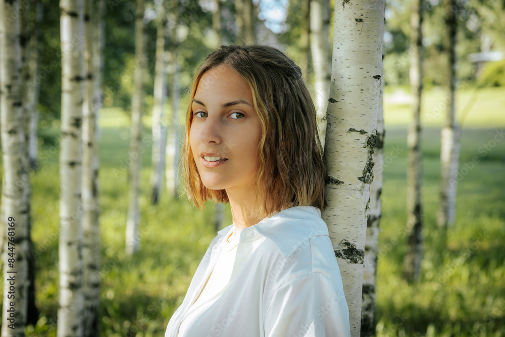 A woman with short hair smiles warmly while leaning against a birch tree in a lush grove. The background is filled with blurred green foliage and trees, creating a serene and natural atmosphere. A woman with short hair smiles warmly while leaning against a birch tree in a lush grove. The background is filled with blurred green foliage and trees, creating a serene and natural atmosphere.