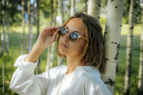 A stylish woman in a white shirt adjusts her sunglasses while standing in a birch tree forest. The sunlight and natural background reflect off her glasses, creating a cool and fashionable look. © Emvats