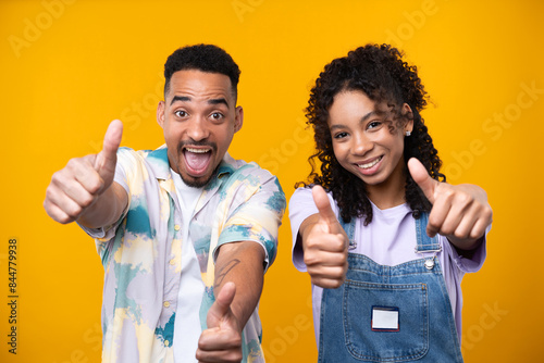 Two hipster friends smiling cheerfully while standing against a yellow background.