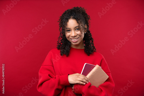 Portrait of a young girl with notebook smiling at the camera while standing against a red background. 