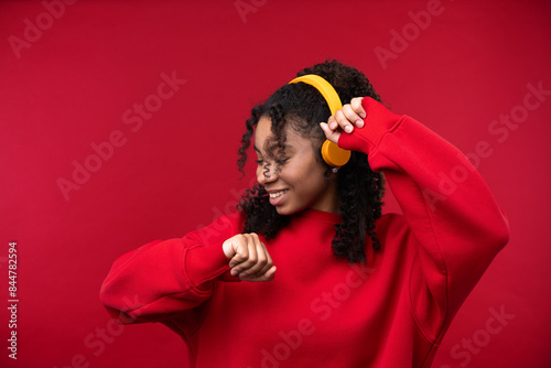 Portrait of a young girl with headphone smiling at the camera while standing against a red background. 