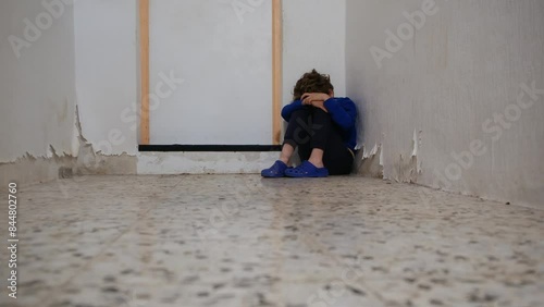 A 6-year-old boy sits alone in a corner of a modest family home, covering his face. Themes include child abuse, isolation, and vulnerability photo