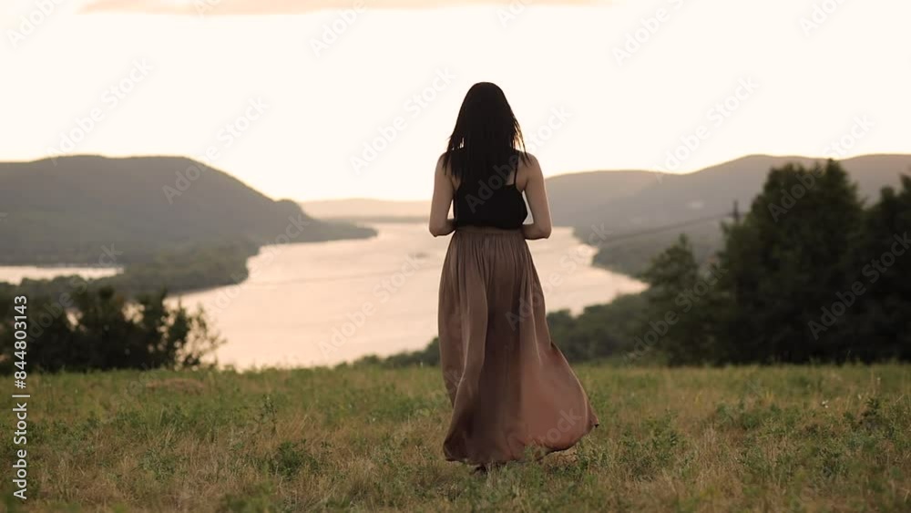 young brunette woman wearing a long skirt dress standing on a field facing away from the camera with mountains and a river in the background in slow motion