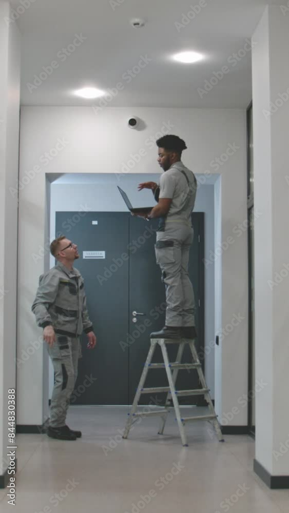 Two multi ethnic installers in uniform sets up security cameras in office corridor using laptop. They talk, check CCTV cameras in computer program. Concept of monitoring system and privacy. Vertical Two multi ethnic installers in uniform sets up security cameras in office corridor using laptop. They talk, check CCTV cameras in computer program. Concept of monitoring system and privacy. Vertical