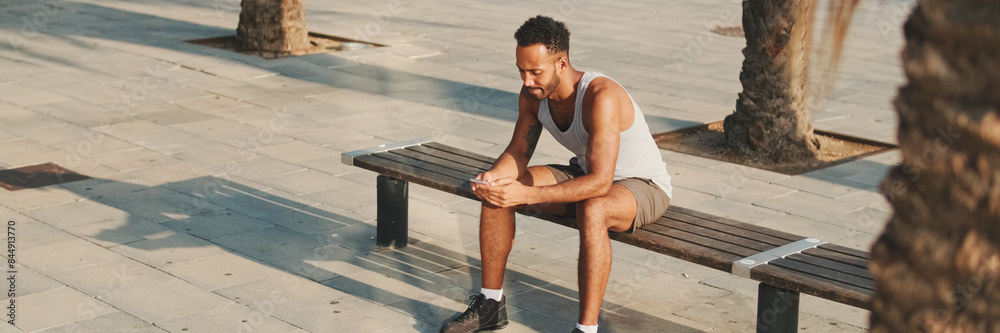 Obraz premium Young bearded male athlete is resting after training, sitting on bench, using smartphone, panorama