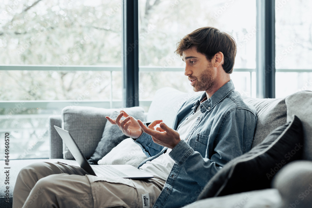 Concentrated 30s man sitting on sofa in living room looking at computer screen, involved in online video call working issues negotiations with colleagues or giving professional consultation to client. Concentrated 30s man sitting on sofa in living room looking at computer screen, involved in online video call working issues negotiations with colleagues or giving professional consultation to client.