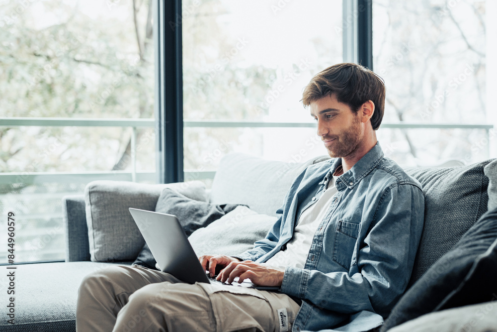 Fototapeta premium Handsome serious man sits on sofa with computer on laps, spend time at home web surfing information, use modern tech, make order on internet. Freelancing, telework, new software learning, tech concept