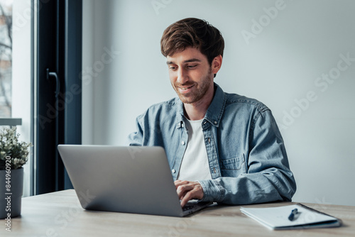 Side view handsome young businessman working with computer remotely, sitting at wooden table in office. Pleasant happy man communicating in social network, searching information online..