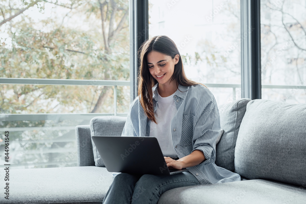 Naklejka premium Concentrated millennial girl sit on couch working on laptop browsing internet at home during lazy weekend, focused young woman freelancer busy using computer surfing wireless web shopping online.