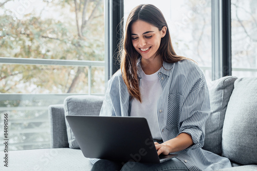 Concentrated millennial girl sit on couch working on laptop browsing internet at home during lazy weekend, focused young woman freelancer busy using computer surfing wireless web shopping online.