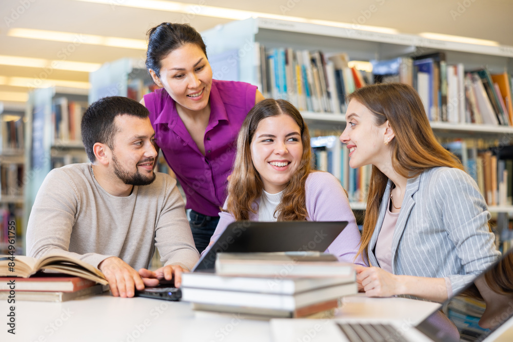 Fototapeta premium Group of positive friendly international university students working on joint research in library, looking for information in books and websites using laptop