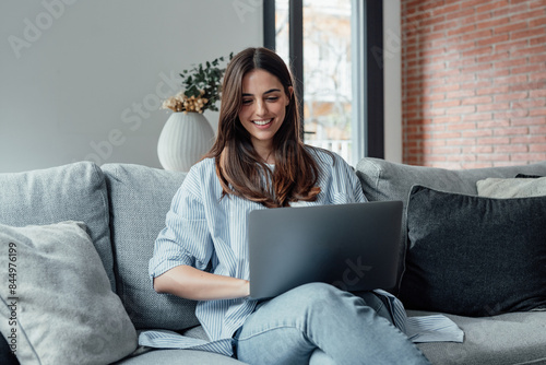 Concentrated millennial girl sit on couch working on laptop browsing internet at home during lazy weekend, focused young woman freelancer busy using computer surfing wireless web shopping online.