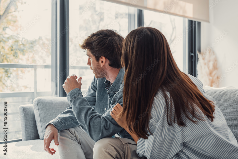 Attractive man is depressed, sitting on the couch ignoring woman, having a dispute with wife or boyfriend, looking desperate and hopeless, having unresolved problem or question. Attractive man is depressed, sitting on the couch ignoring woman, having a dispute with wife or boyfriend, looking desperate and hopeless, having unresolved problem or question.