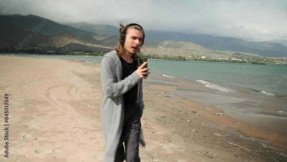 Young guy in casual clothes walks along the beach and sings along to the music playing on his phone, headphones, relaxing on the weekend. Young guy in casual clothes walks along the beach and sings along to the music playing on his phone, headphones, relaxing on the weekend.
