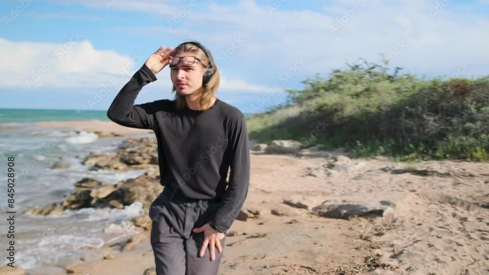 Cool young guy in black clothes flirting to music on headphones while walking along the sandy beach on a summer day. Cool young guy in black clothes flirting to music on headphones while walking along the sandy beach on a summer day.
