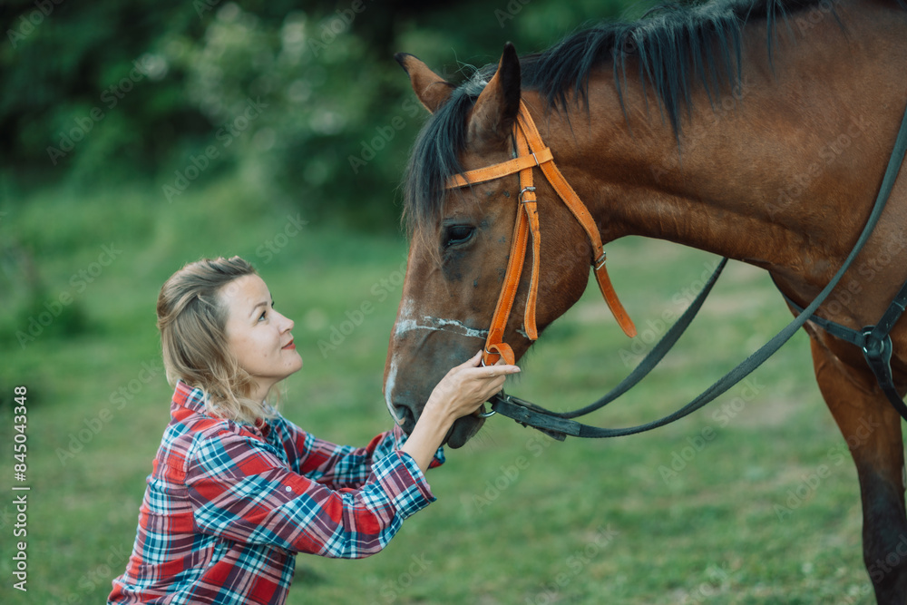 Obraz premium Happy blonde with horse in forest. Woman and a horse walking through the field during the day. Dressed in a plaid shirt and black leggings.
