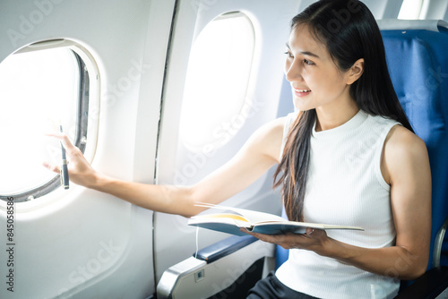 A young Asian woman, an airplane passenger, sits by the window seat, studying abroad. She gazes out the window, ready for takeoff in the economy class section. photo