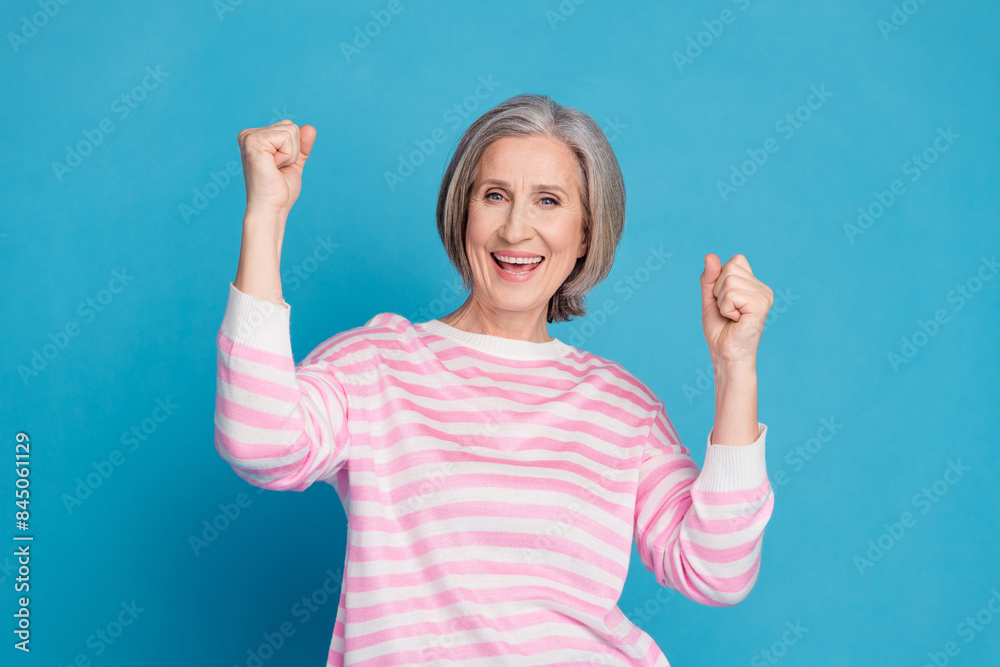 Photo portrait of attractive retired woman raise fists winning dressed stylish striped clothes isolated on blue color background Photo portrait of attractive retired woman raise fists winning dressed stylish striped clothes isolated on blue color background