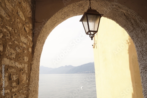 Fototapeta Naklejka Na Ścianę i Meble -  View of a glimpse of Lake Como from a quaint corner of an alley in the historic center of Varenna that directly overlooks the water of Lake. Varenna , Lake Como, Italy, village, street, street lamp,