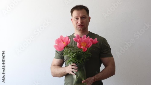 A man in a hacky t-shirt holds a vase of peonies and points to the camera photo