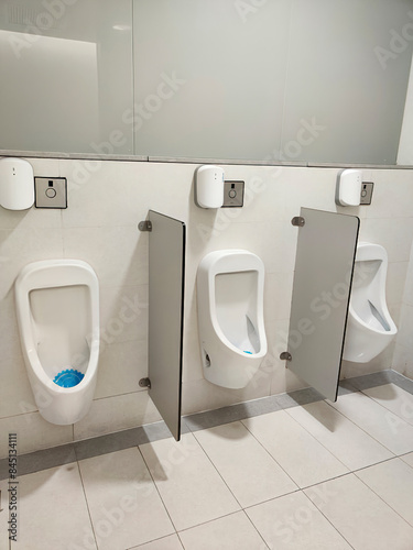 A row of white urinals in a public toilet against a tiled wall. Each urinal is separated by minimal partitions for privacy, with a clean and well-maintained environment.