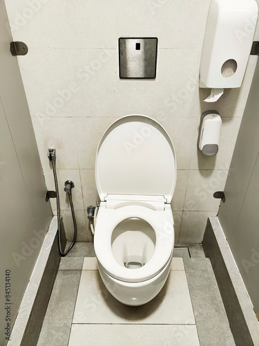 Inside a public toilet cubicle, white walls surround a toilet with a closed lid. A toilet paper roll holder is visible, and the floor is clean. Privacy is maintained by a locked door.