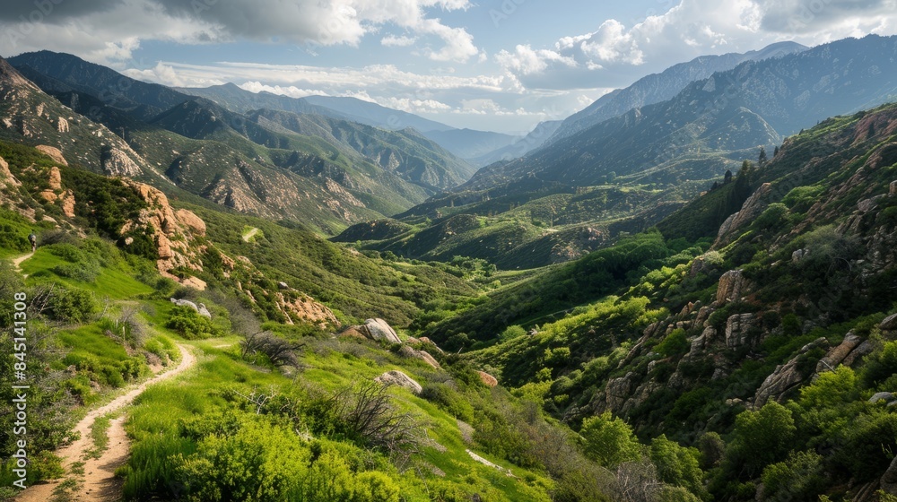 A wide-angle view of a winding path leading down through a lush green valley surrounded by rugged mountains. The sky is cloudy, hinting at a potential storm