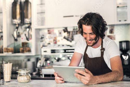 Handsome owner of coffee shop, barista leaning on counter and making order from supplier on tablet.