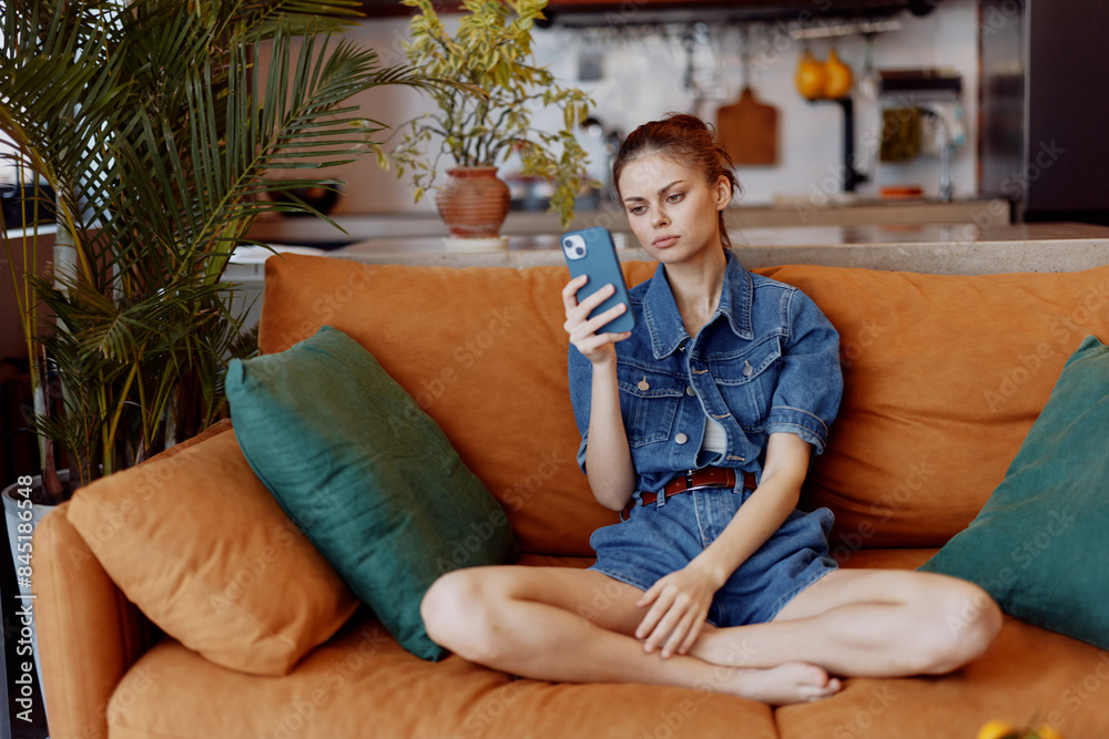 Obraz premium Attractive woman sitting on orange couch, holding cell phone and absorbed in digital device